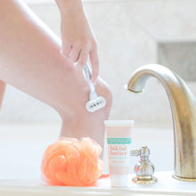 Luxe Shaving Cream Bikini Cutter scented with Mixologie Tempted (Coconut Kiss) Fragrance in a 2 fl oz or 60 mL white tube with orange circle pattern and white cap. Bikini Butter pictured on a bathtub with loofah while model shaves leg in background.  